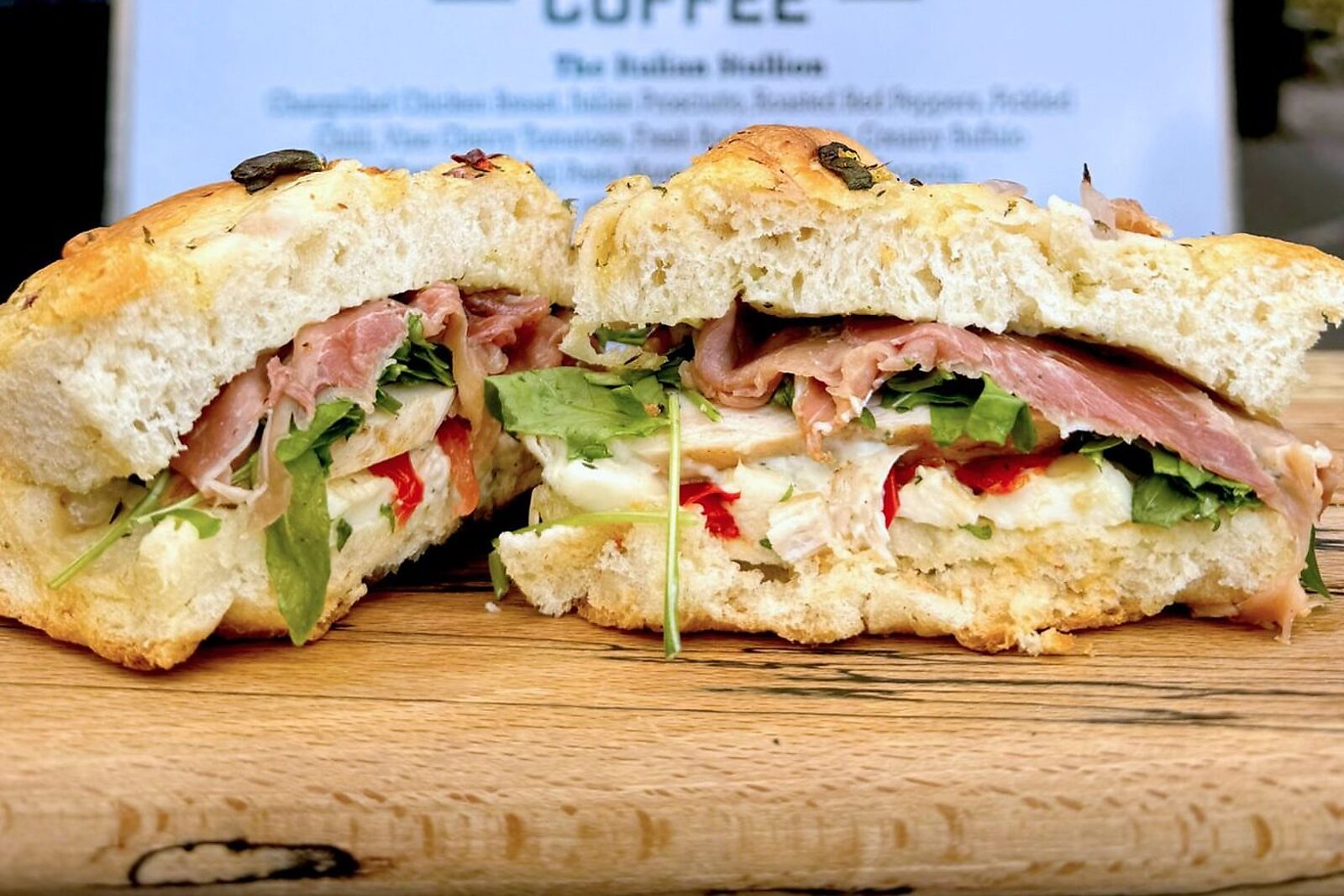 A focaccia sandwich cut in half on a wooden board, with the Box'd Coffee sign behind it