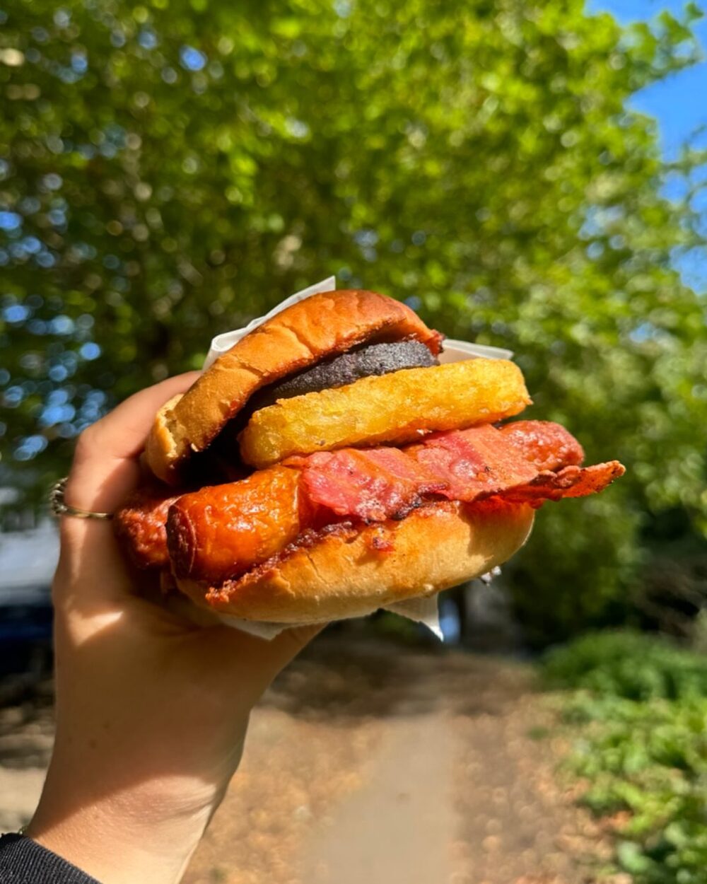 A hand holding a breakfast burger outdoors under trees